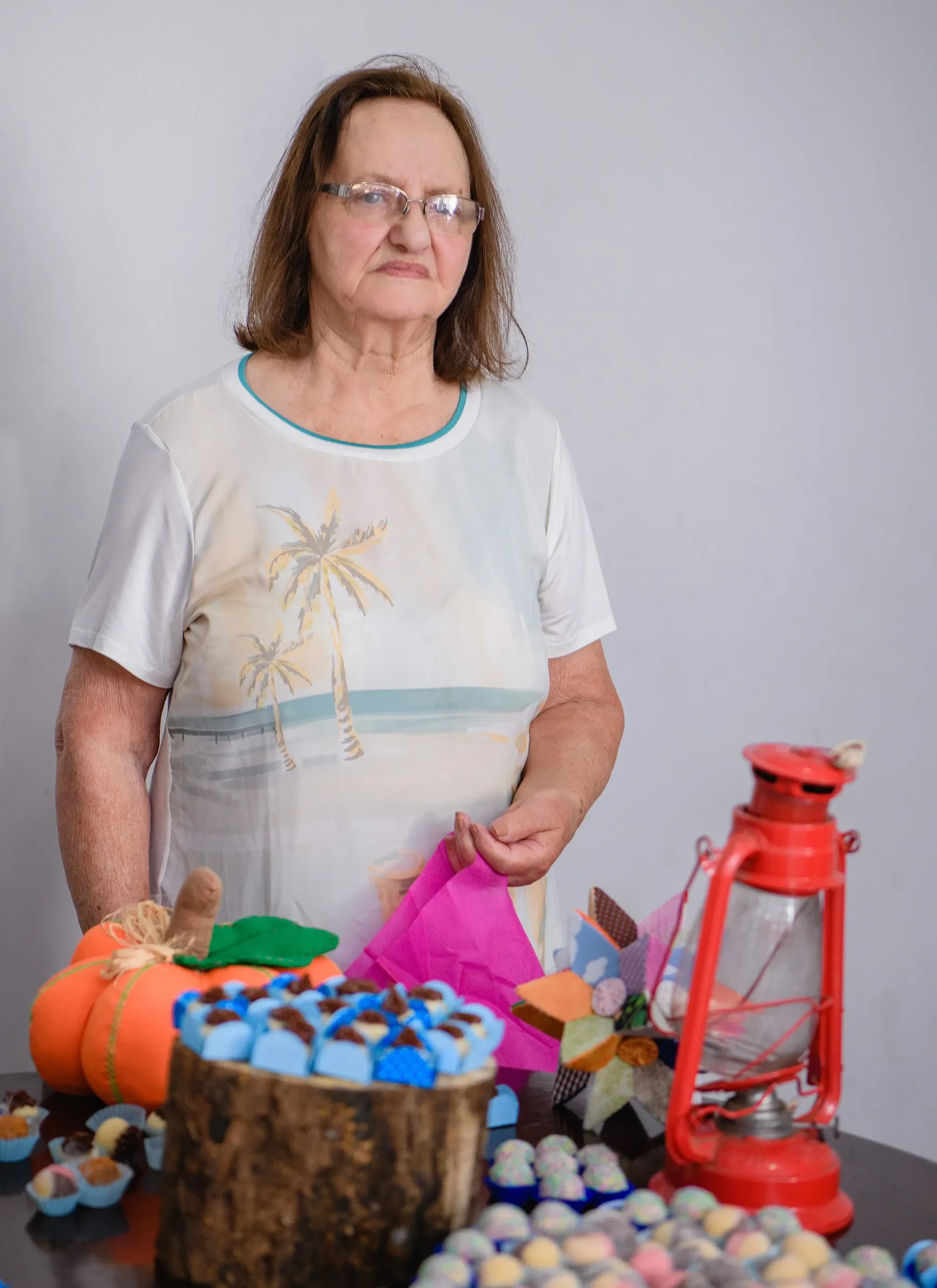 Mulher de óculos, com uma blusa branca estampada com palmeiras, olha para a direita enquanto está atrás de uma mesa decorada com um bolo azul, abóbora de feltro e uma lanterna vermelha.