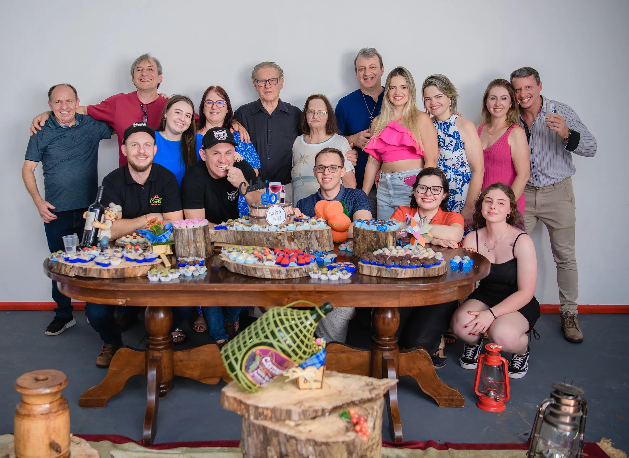 Um grupo heterogêneo de 19 pessoas, de diversas idades, está posando atrás de uma mesa de madeira decorada com vários doces, bolos e garrafas de vinho, com uma parede branca lisa ao fundo.