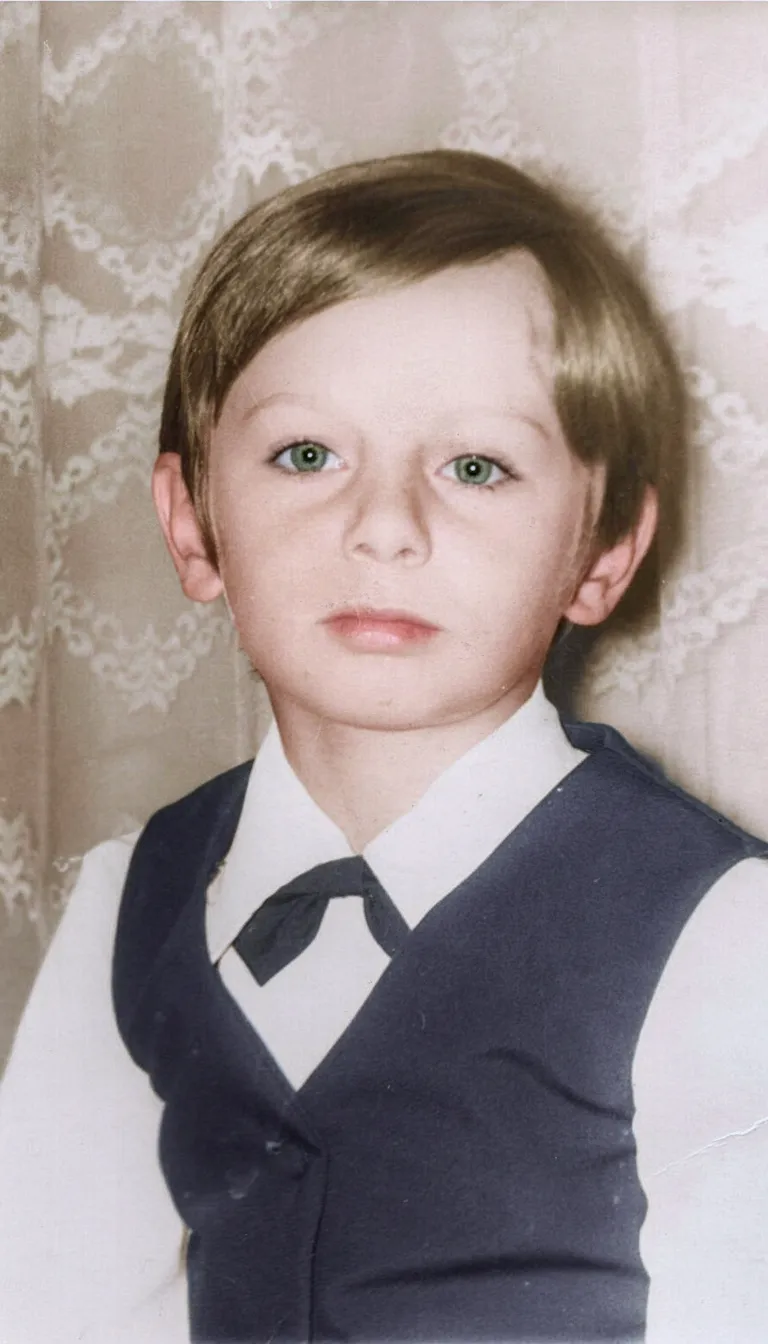 Um menino com cabelo castanho e olhos verdes, vestindo uma camisa branca, um colete azul escuro e uma gravata borboleta, olhando diretamente para a câmera.