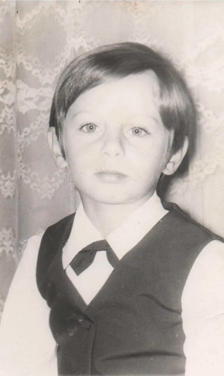 Um menino com cabelo castanho e olhos verdes, vestindo uma camisa branca, um colete azul escuro e uma gravata borboleta, olhando diretamente para a câmera. (Original)