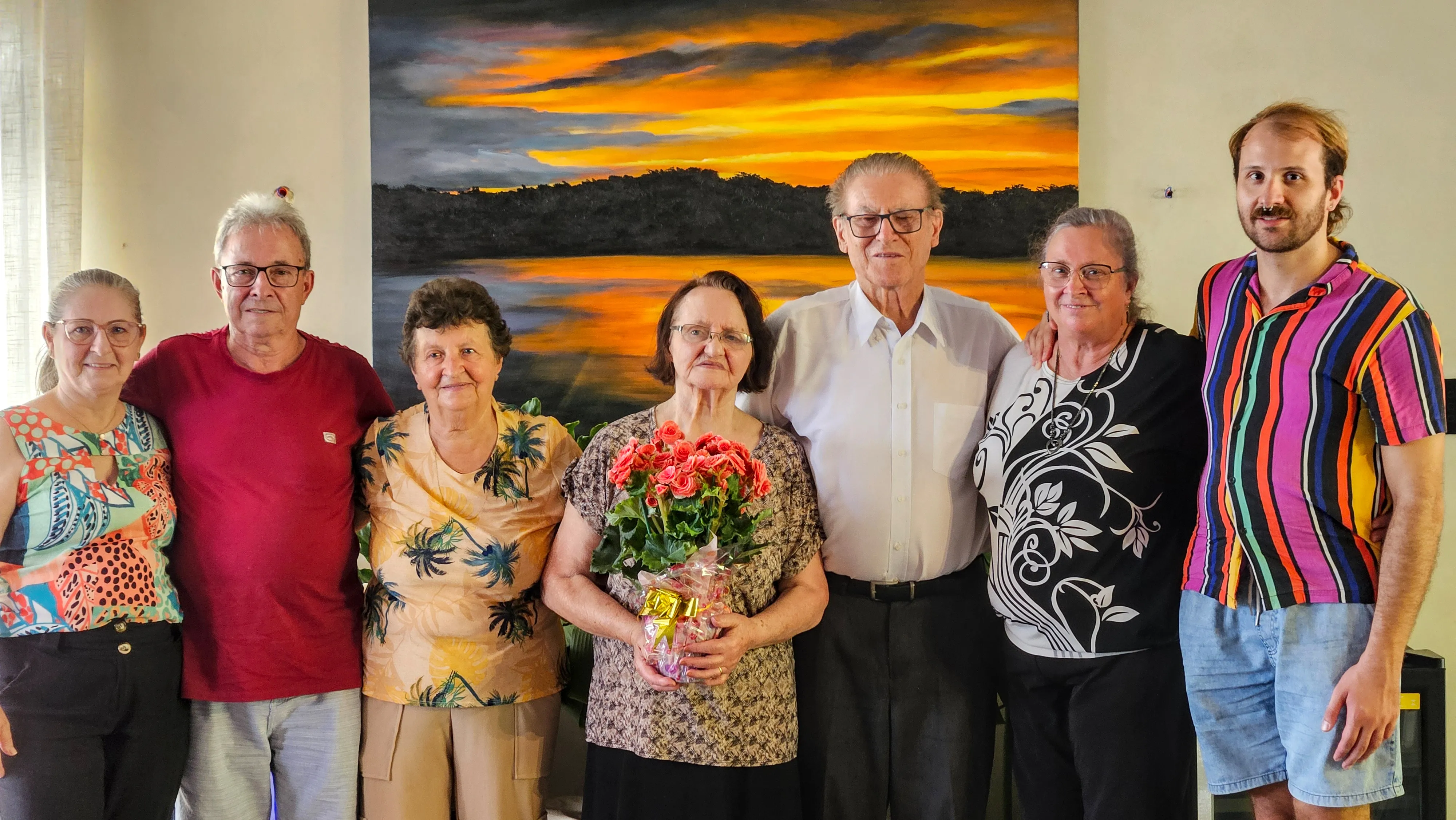 Seis pessoas, incluindo dois idosos no centro, posam para uma foto em frente a uma pintura de pôr do sol, com uma mulher segurando um buquê de flores.