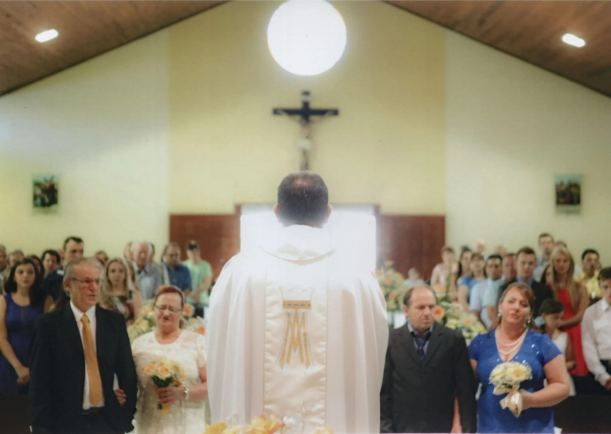 Um padre, de costas para a câmera, está celebrando uma missa em uma igreja cheia, com o altar e um crucifixo ao fundo.