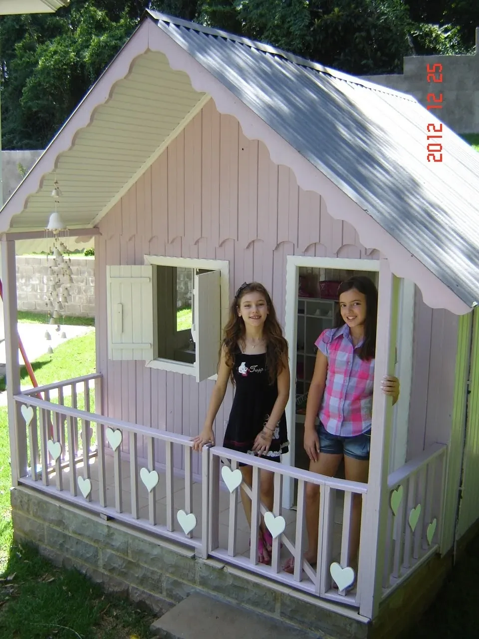 Duas meninas sorriem na varanda de uma casa de brinquedo de madeira rosa com telhado de metal cinza.