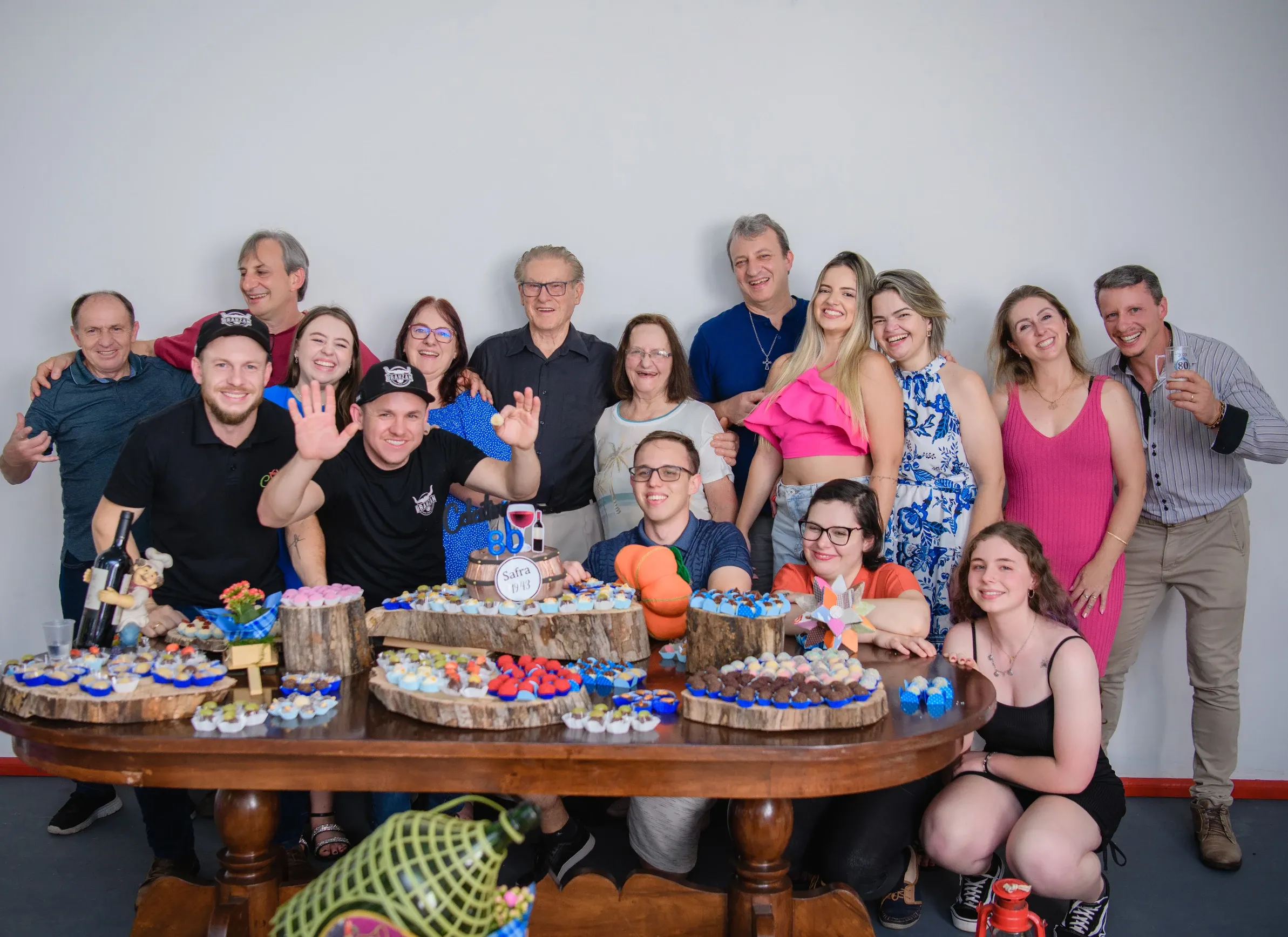 Um grande grupo de pessoas sorridentes, de várias idades, posando atrás e em volta de uma mesa de madeira cheia de doces e garrafas de vinho, com alguns fazendo gestos de "paz" ou acenando.