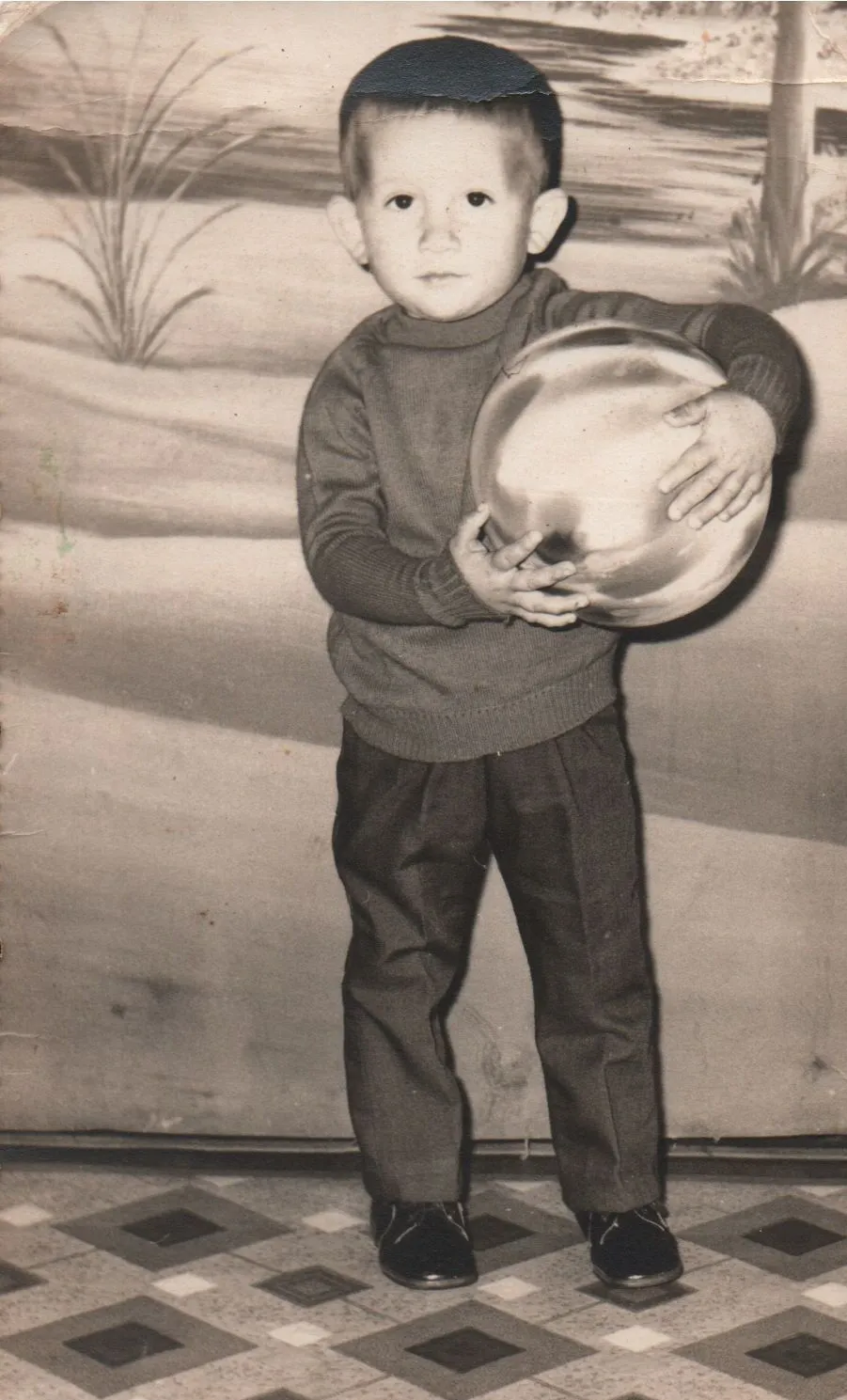 Um menino pequeno de camisa polo cor de vinho e calças cinzas segura uma bola prateada e cinza contra o peito, em pé em um chão de azulejos na frente de um pano de fundo bege com uma leve pintura de grama. (Original)