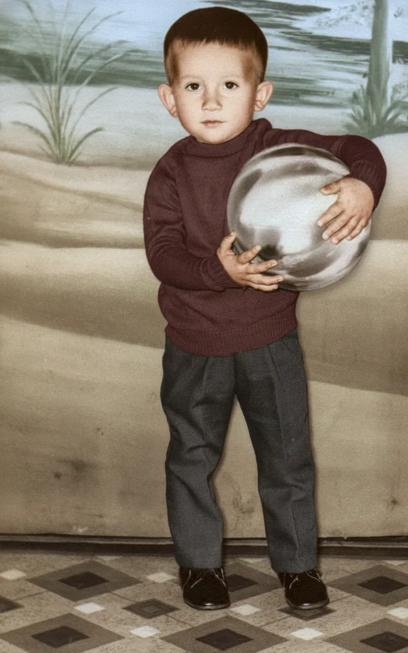 Um menino pequeno de camisa polo cor de vinho e calças cinzas segura uma bola prateada e cinza contra o peito, em pé em um chão de azulejos na frente de um pano de fundo bege com uma leve pintura de grama.