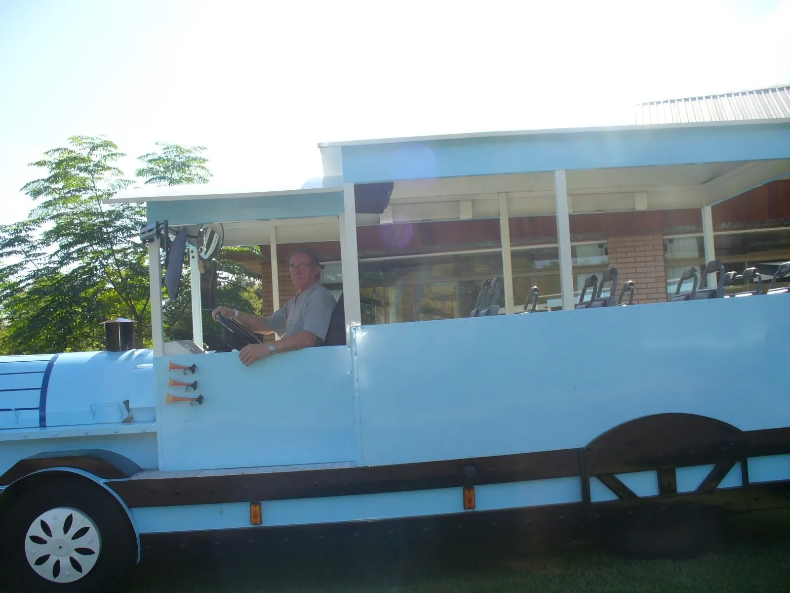 Homem de óculos dirigindo um grande veículo aquático azul e branco, em um dia ensolarado, com árvores e um prédio no fundo.