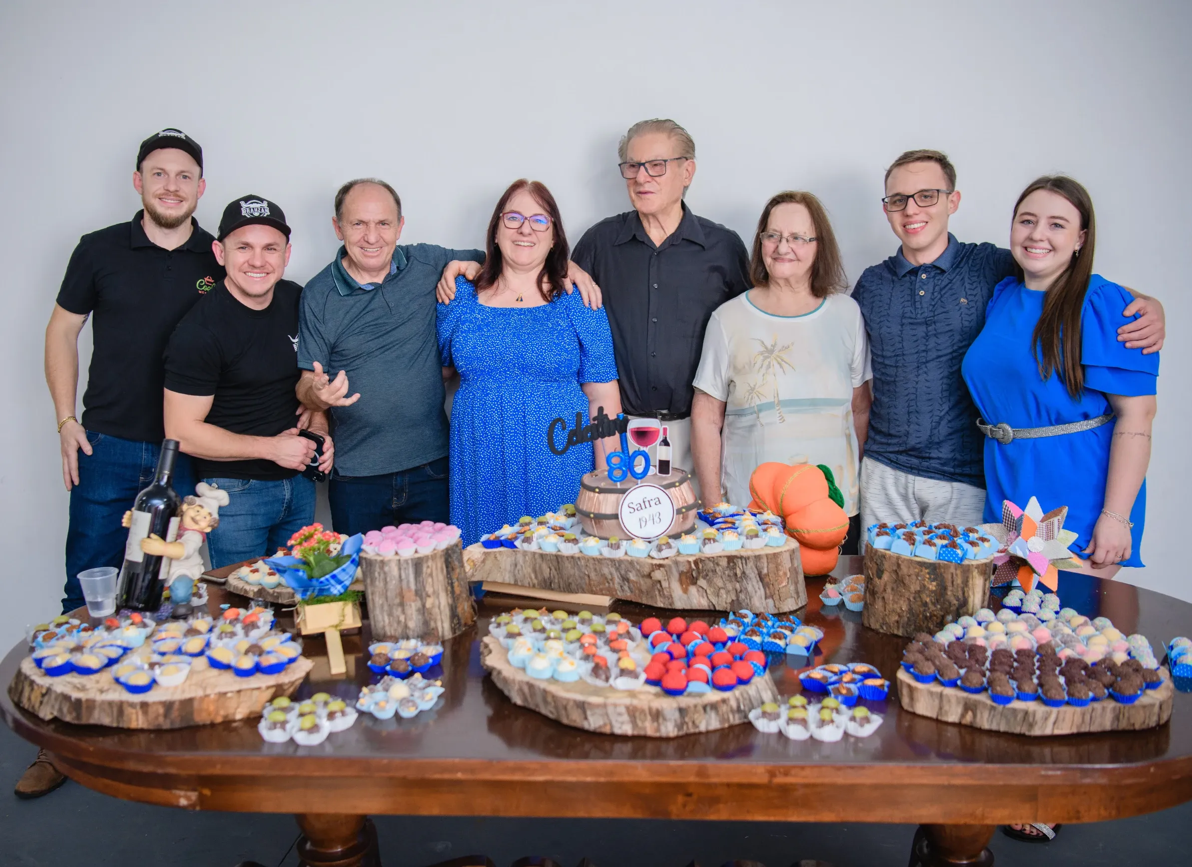 Um grupo de oito pessoas sorri atrás de uma mesa cheia de doces e um bolo de aniversário, celebrando.