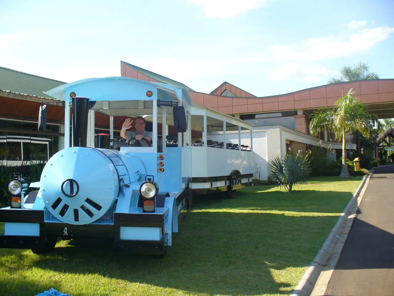 Homem de meia-idade acena do banco do motorista de um pequeno trem turístico azul-claro com um trailer branco, em um gramado ao lado de um caminho pavimentado e prédios modernos com telhados angulares.