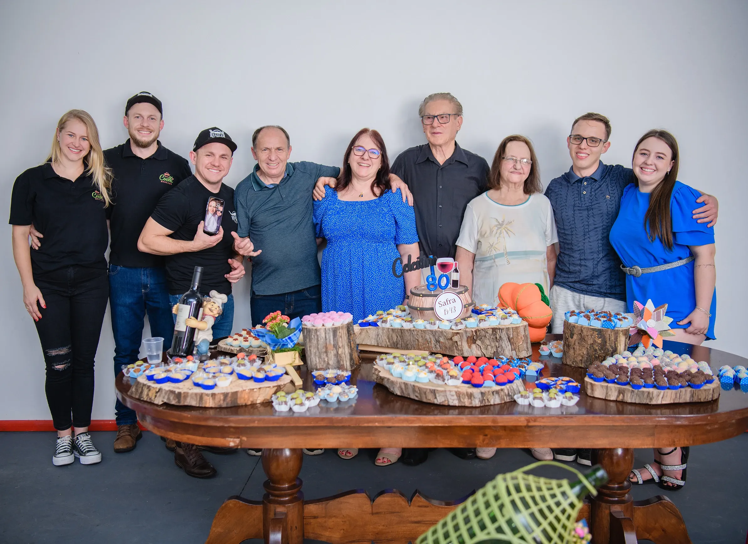 Um grupo de dez pessoas (cinco homens e cinco mulheres) posa para uma foto de família em frente a uma mesa com um bolo e vários doces.