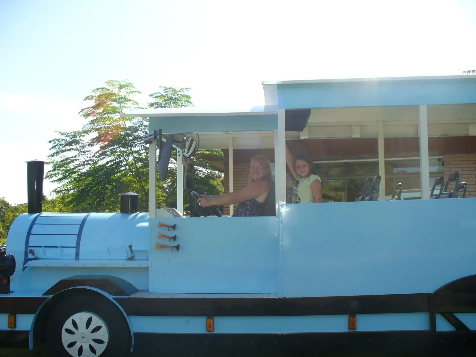 Uma mulher e uma garota sorridentes sentam na frente de um trem azul com janelas, a mulher no banco do motorista e a garota acenando com a mão.