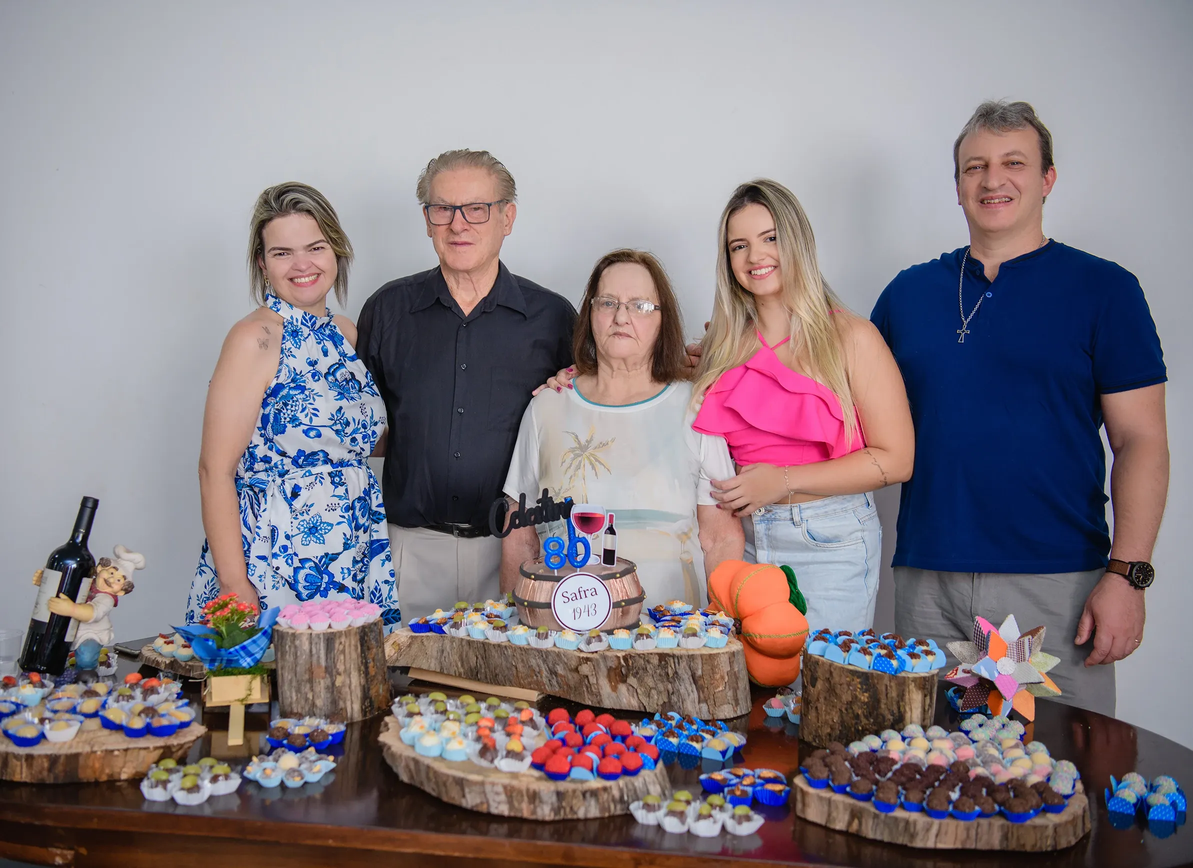 Cinco pessoas sorriem para a câmera atrás de uma mesa com vários doces e um bolo de aniversário de 80 anos.