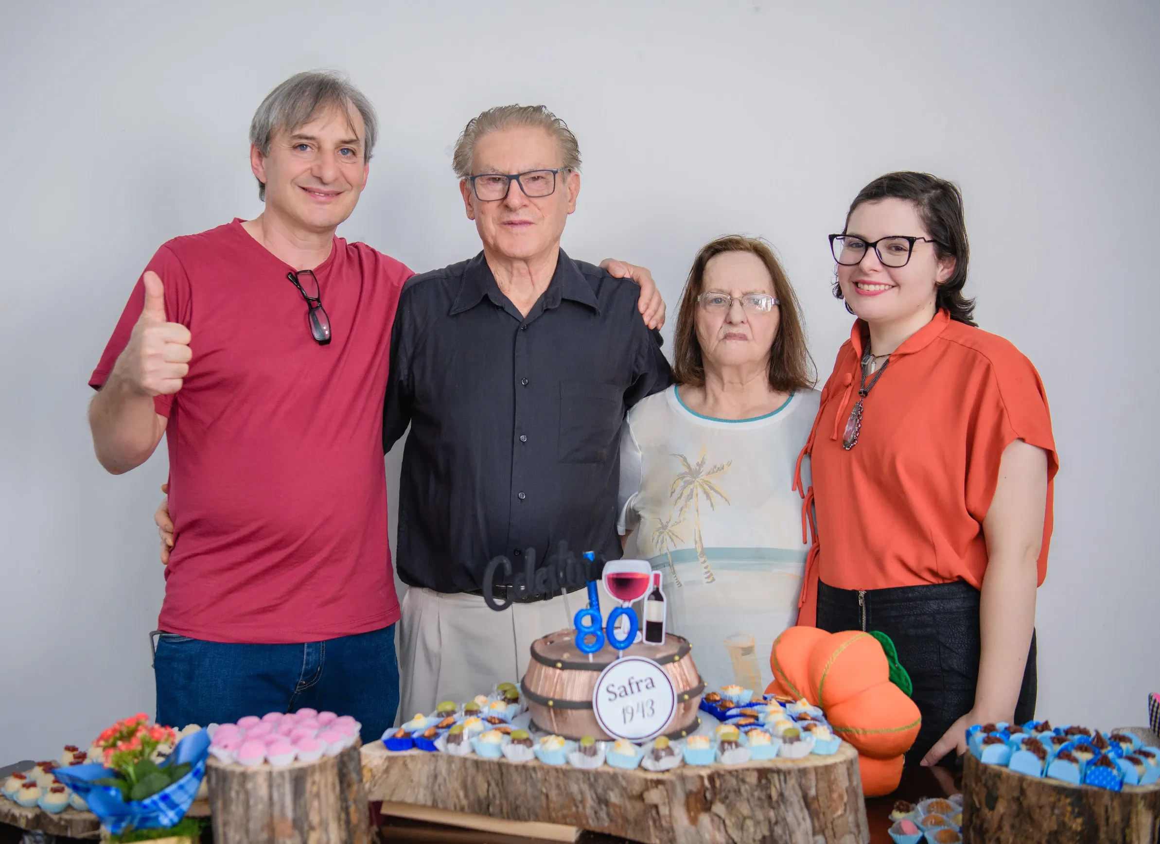 Quatro pessoas, duas mulheres e dois homens, sorriem para a câmera em uma celebração de aniversário, com um bolo e doces em uma mesa à frente.