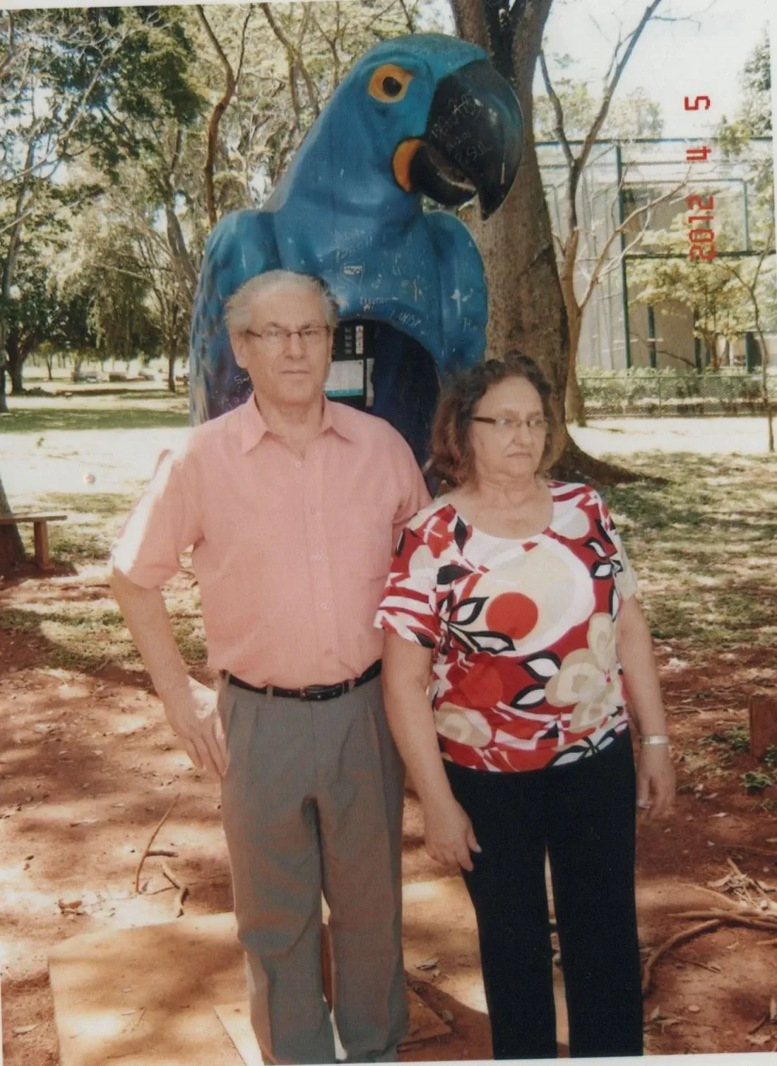Um casal de idosos de pé em um parque, com um homem de camisa rosa e calças cinzas à esquerda e uma mulher de blusa estampada e calças pretas à direita. Atrás deles, há uma grande estátua de papagaio azul.