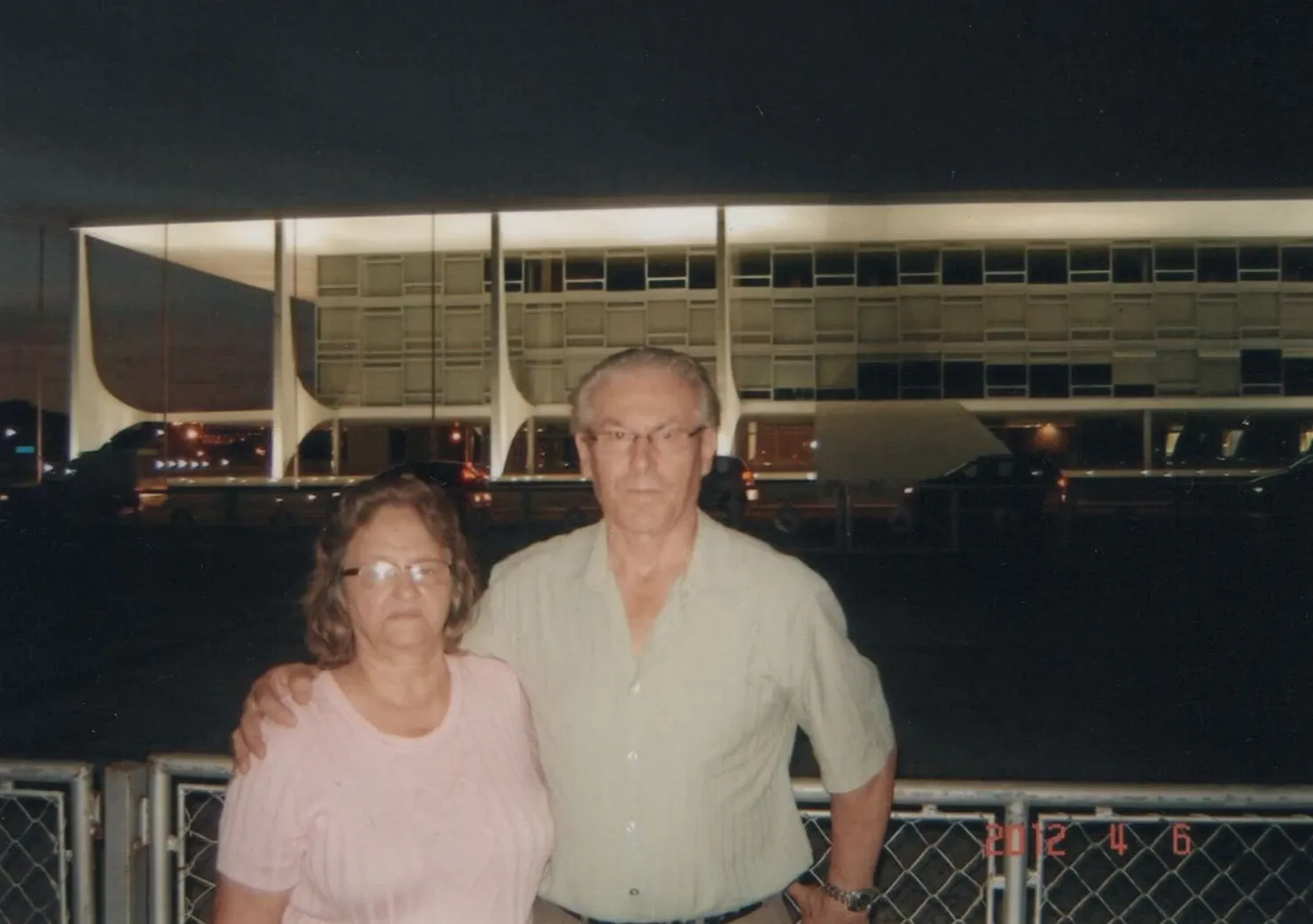 Casal de meia-idade posa à noite em frente a um prédio moderno iluminado com muitas janelas.