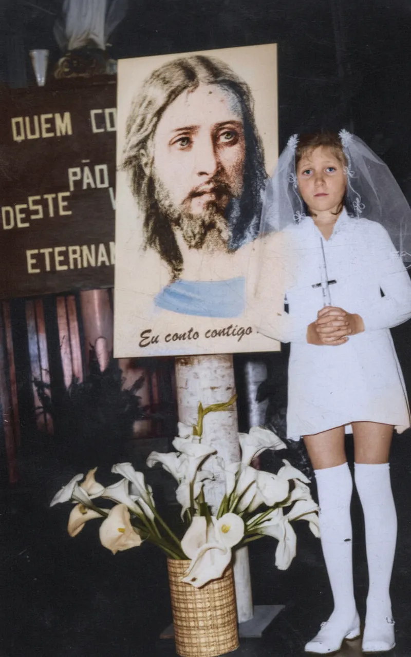 Menina em primeira comunhão em pé ao lado de um retrato de Jesus Cristo, segurando um crucifixo e usando um vestido branco, meias brancas e sapatos brancos com um véu na cabeça, com um vaso de lírios brancos aos seus pés.