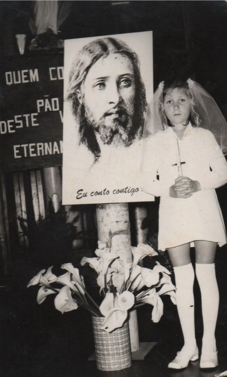 Menina em primeira comunhão em pé ao lado de um retrato de Jesus Cristo, segurando um crucifixo e usando um vestido branco, meias brancas e sapatos brancos com um véu na cabeça, com um vaso de lírios brancos aos seus pés. (Original)