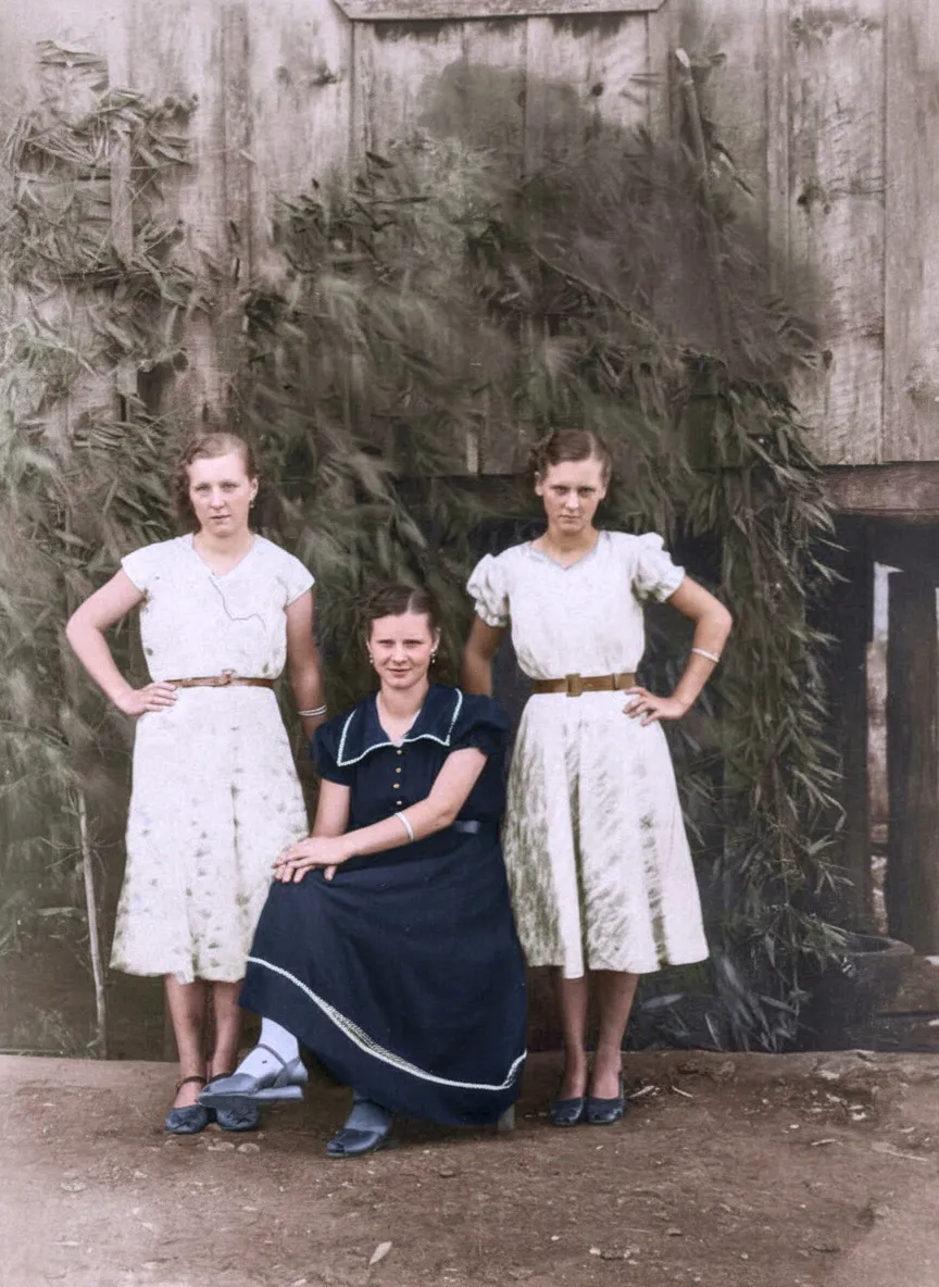 Três mulheres posam em frente a uma parede de madeira; duas estão de pé usando vestidos brancos, e uma está sentada usando um vestido azul-marinho.