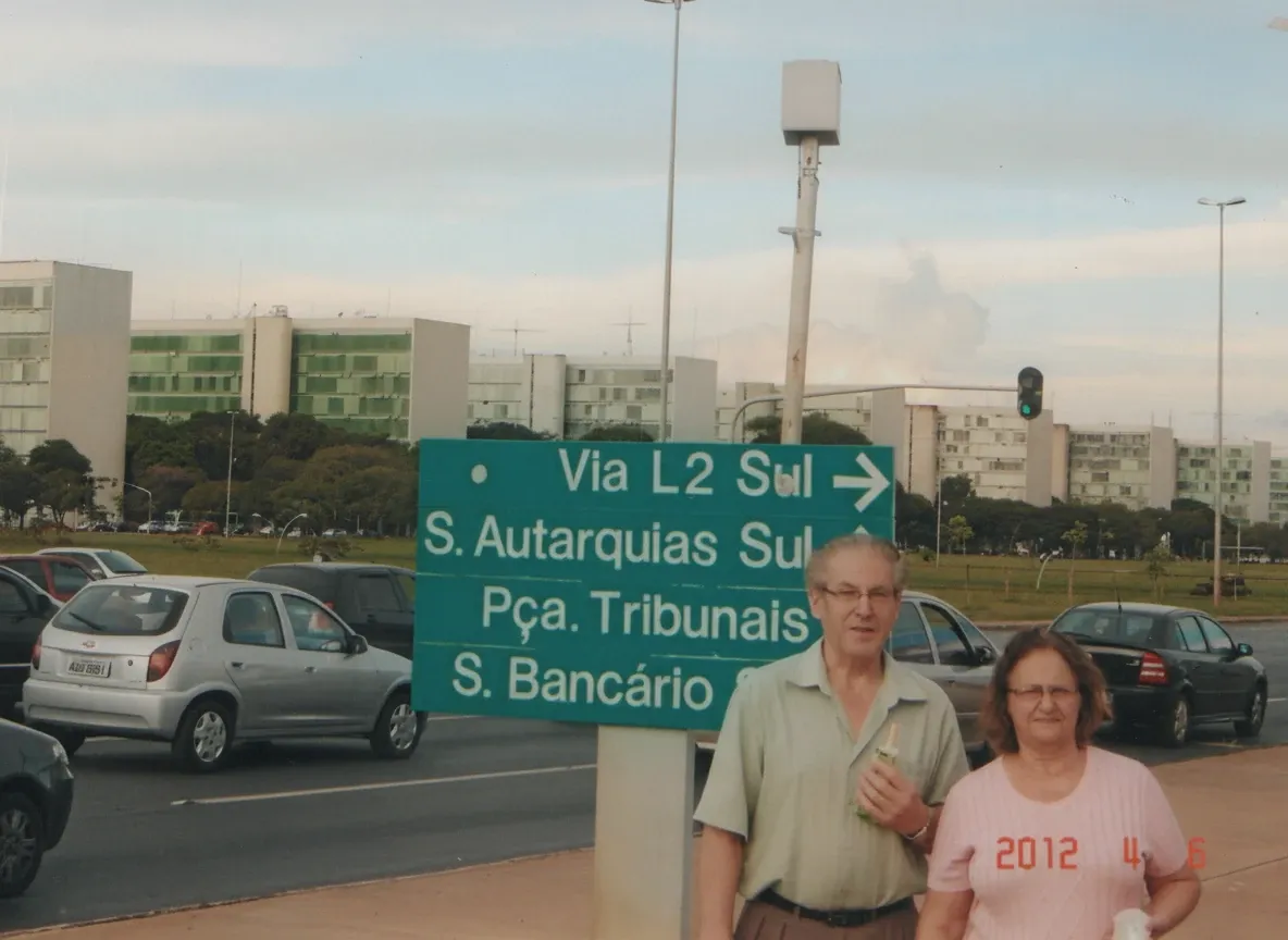 Um homem idoso e uma mulher em pé na beira de uma rua, com carros e edifícios ao fundo, e uma placa de rua verde que aponta para a "Via L2 Sul".