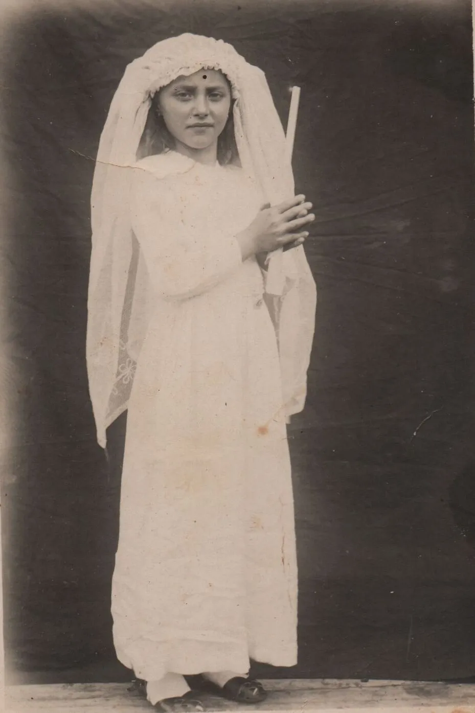 Jovem de cabelo ruivo usando vestido de primeira comunhão branco com véu e segurando uma vela em pé. (Original)