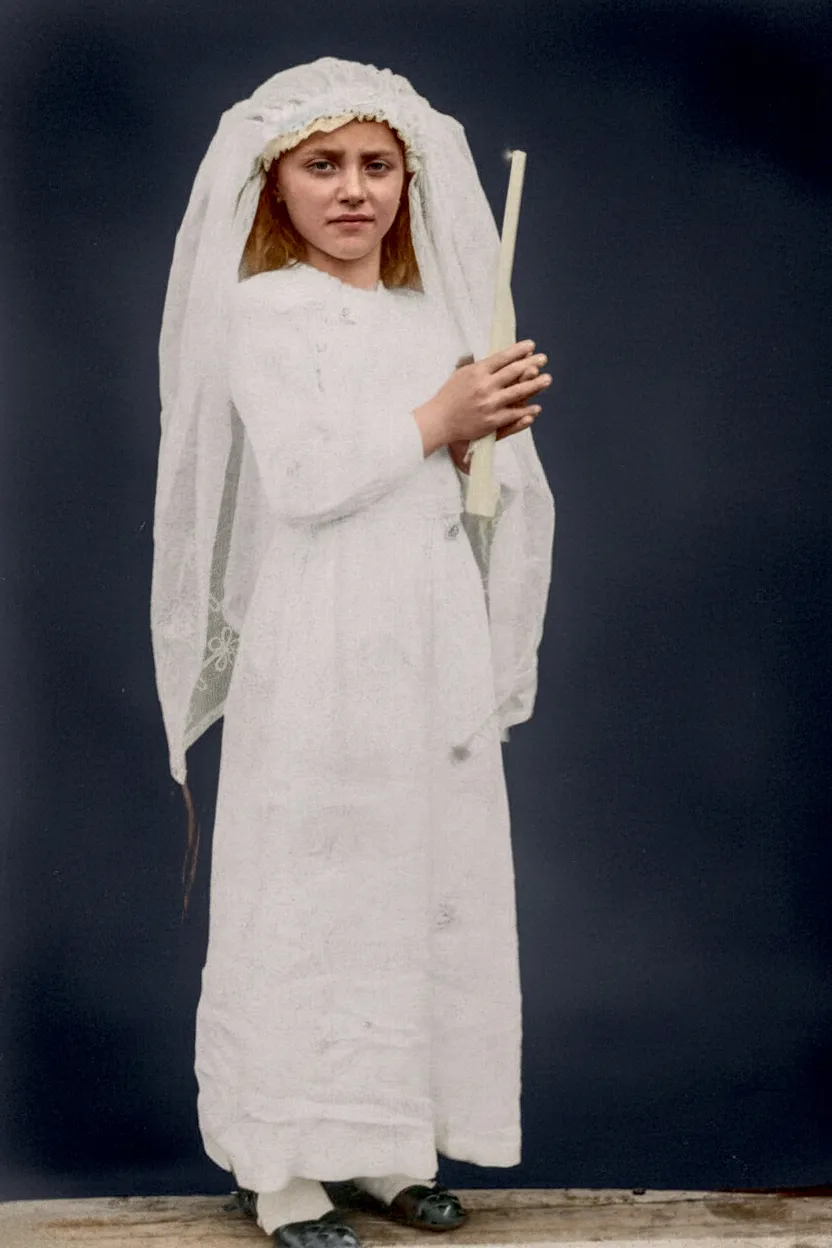 Jovem de cabelo ruivo usando vestido de primeira comunhão branco com véu e segurando uma vela em pé.