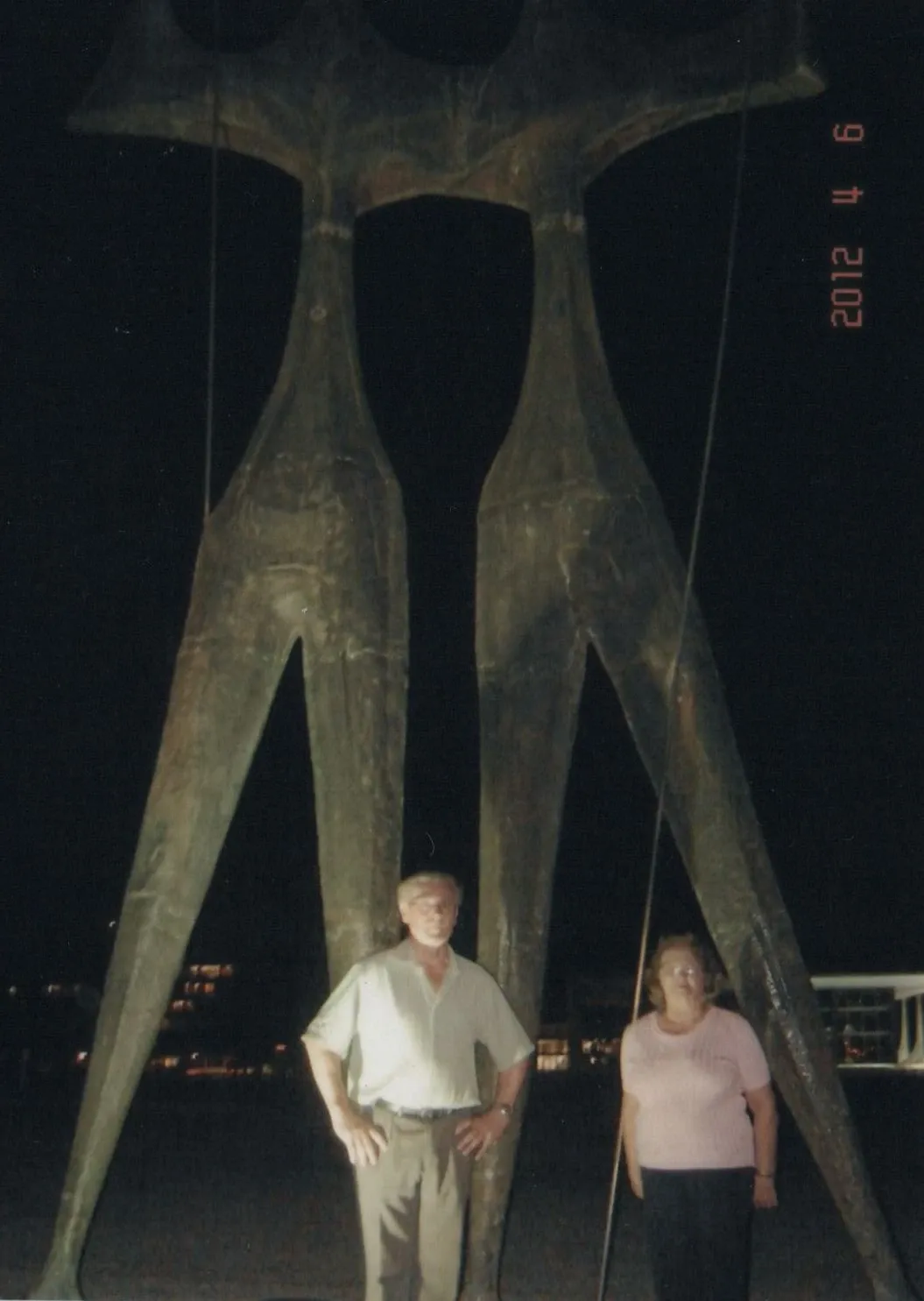 Um homem e uma mulher, vistos da cintura para cima, em frente a uma enorme escultura abstrata escura de duas figuras alongadas e esguias em um ambiente noturno com pouca iluminação.