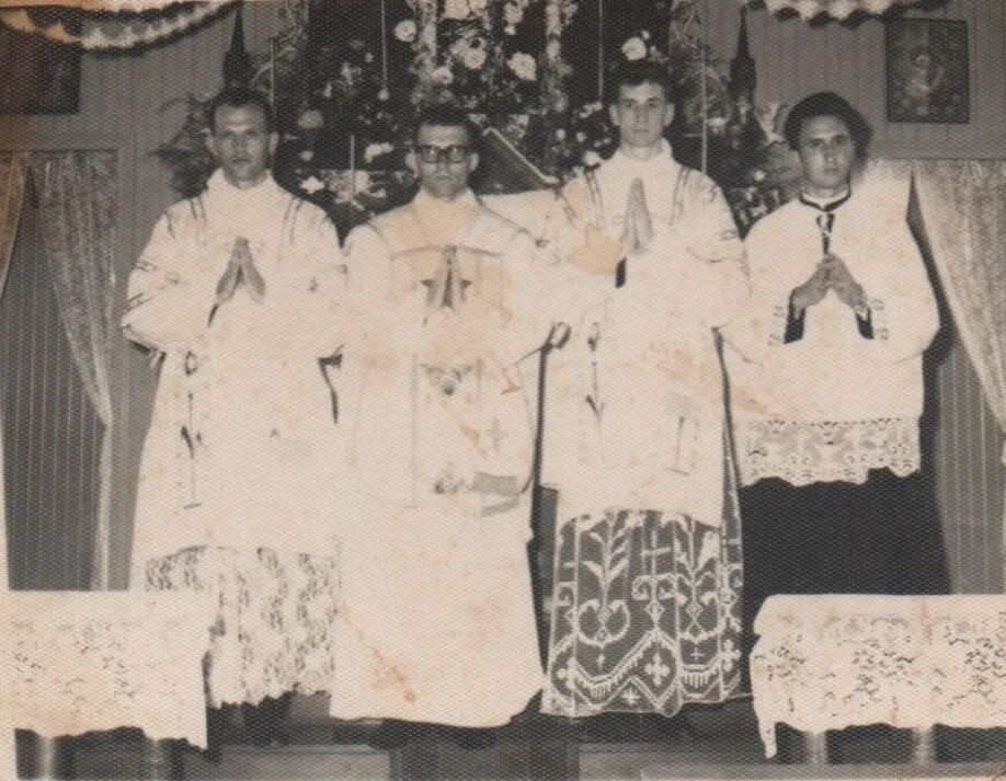 Quatro padres estão de pé na frente de um altar floral, com as mãos cruzadas em oração. (Original)