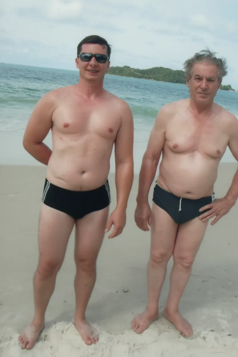 Dois homens, um mais jovem com óculos de sol e o outro mais velho, em fatos de banho na areia de uma praia com o oceano e ilhas ao fundo.