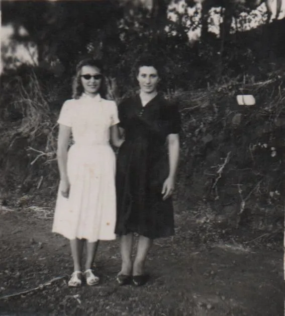 Duas mulheres em pé em um caminho de terra com folhagem atrás delas; a mulher à esquerda veste um vestido branco e óculos de sol e a mulher à direita veste um vestido azul marinho. (Original)