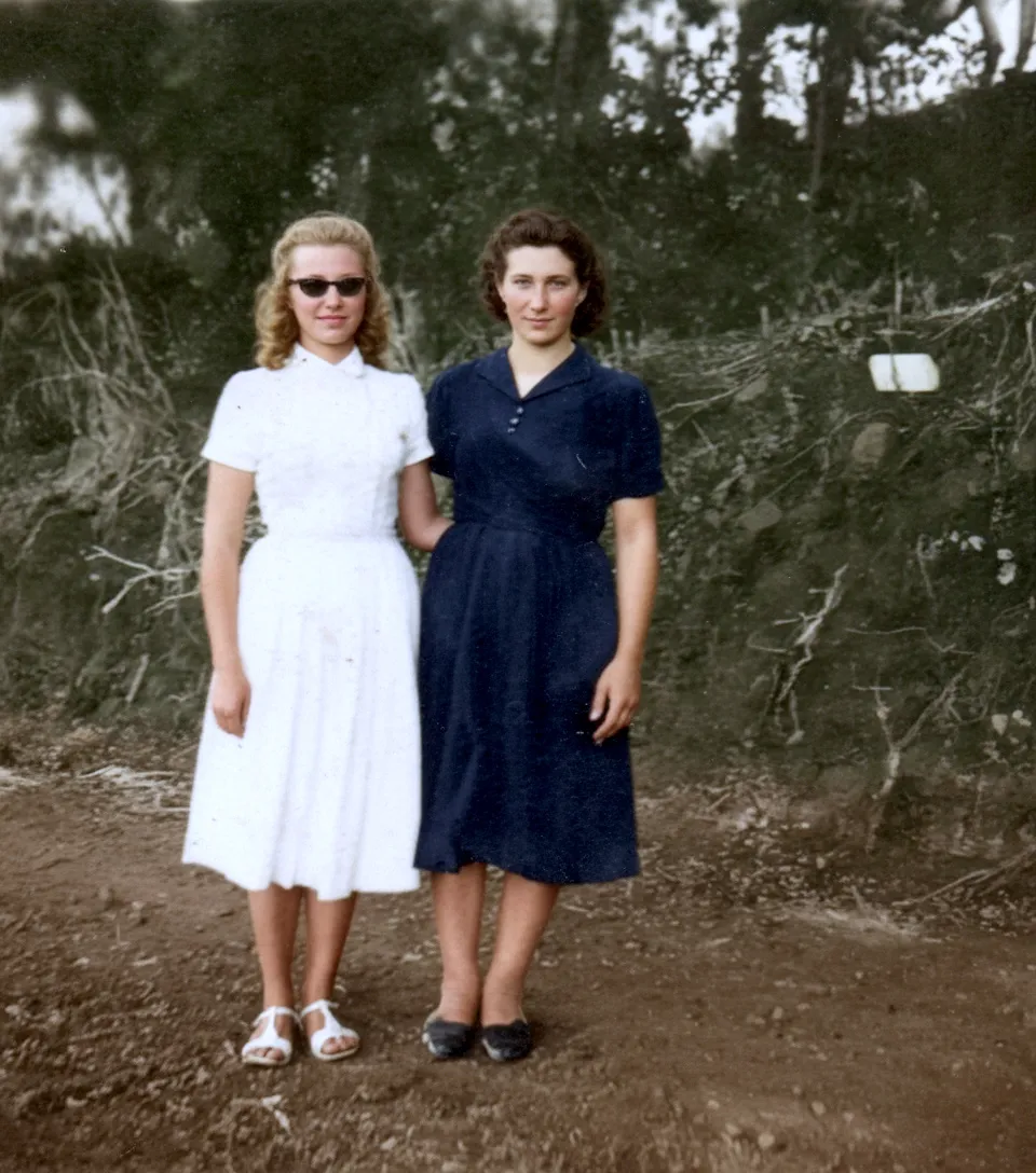 Duas mulheres em pé em um caminho de terra com folhagem atrás delas; a mulher à esquerda veste um vestido branco e óculos de sol e a mulher à direita veste um vestido azul marinho.