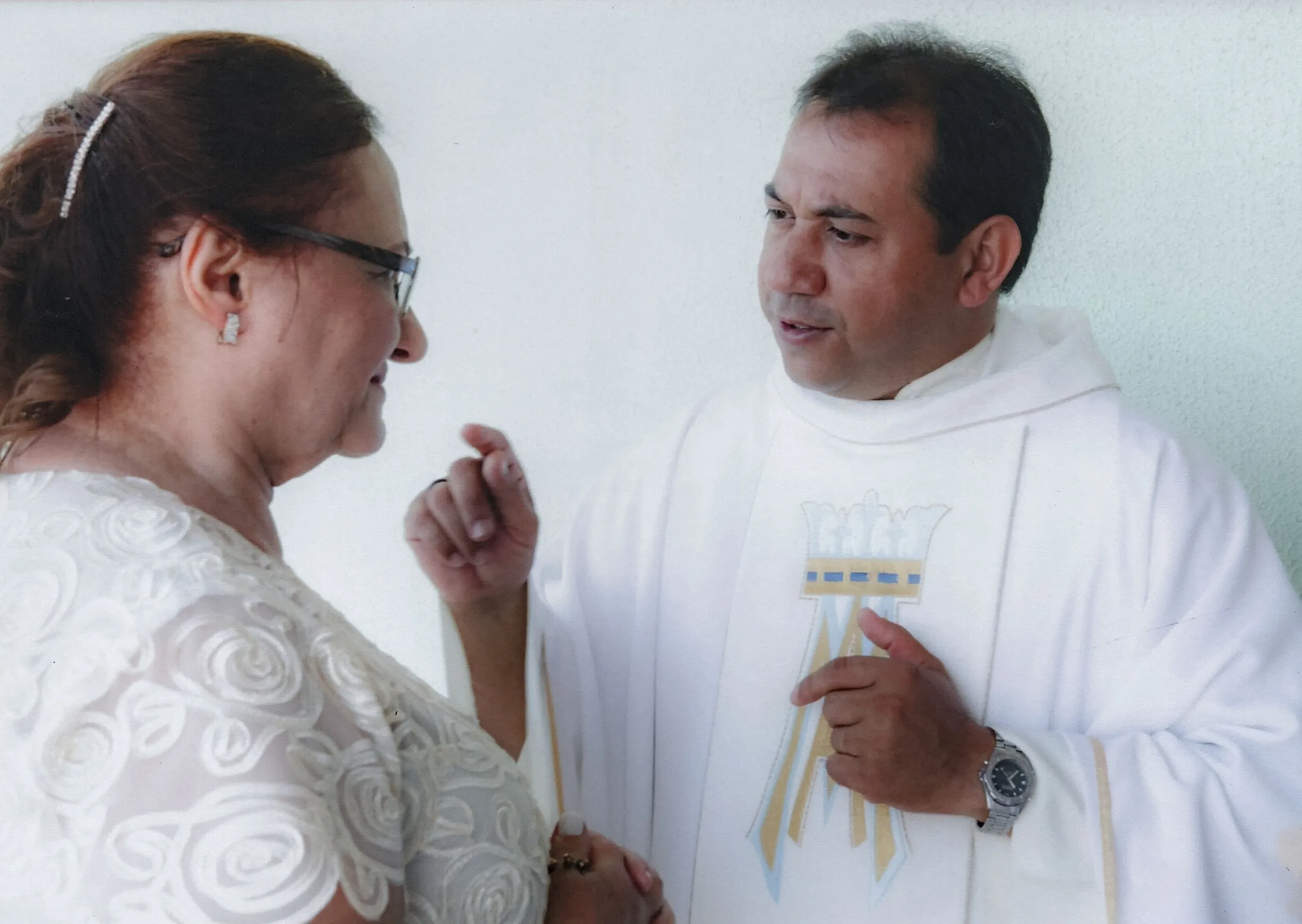 Um padre de meia-idade em vestes brancas olha e gesticula para uma mulher de meia-idade de óculos e vestido branco, que olha para ele com um sorriso.
