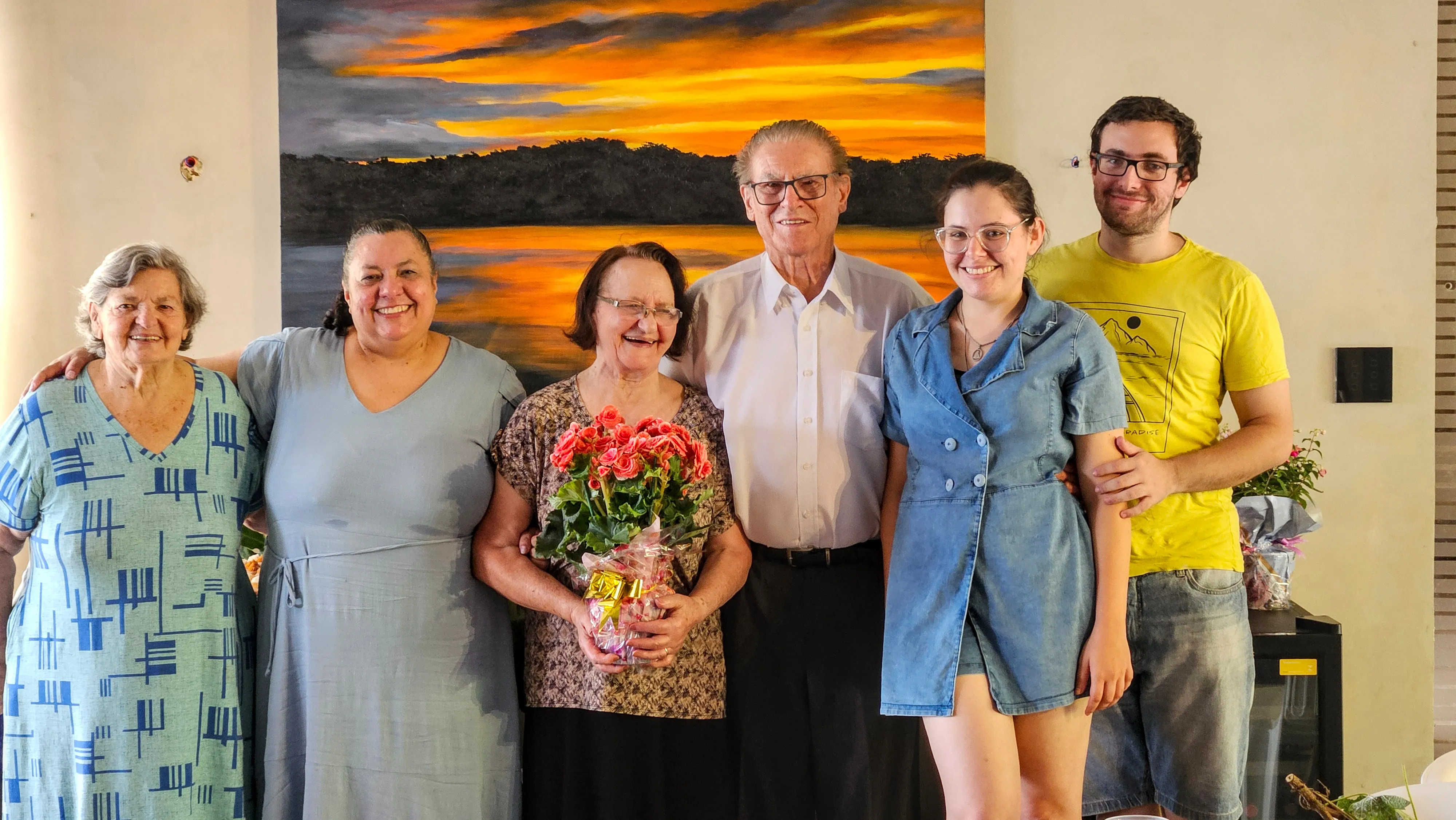 Um grupo de seis pessoas, três mulheres e três homens de diferentes idades, está posando em frente a uma grande pintura de um pôr do sol colorido sobre uma paisagem escura; a mulher no centro segura um buquê de flores vermelhas.