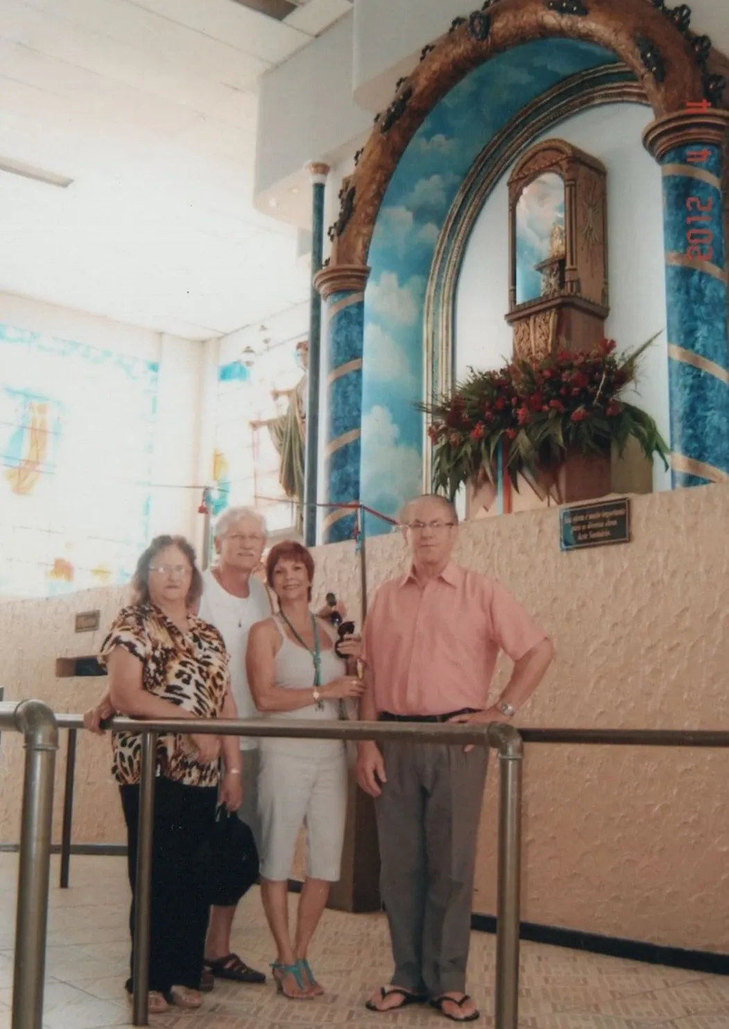 Quatro pessoas, duas mulheres e dois homens, posam em um templo iluminado, com a mulher à esquerda e o homem à direita apoiados em um corrimão, enquanto os outros dois ficam entre eles.