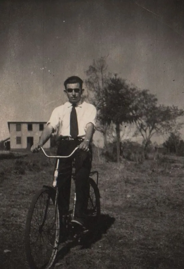 Homem de óculos escuros, camisa de manga curta e gravata azul, em pé ao lado de uma bicicleta preta em um campo gramado. (Original)