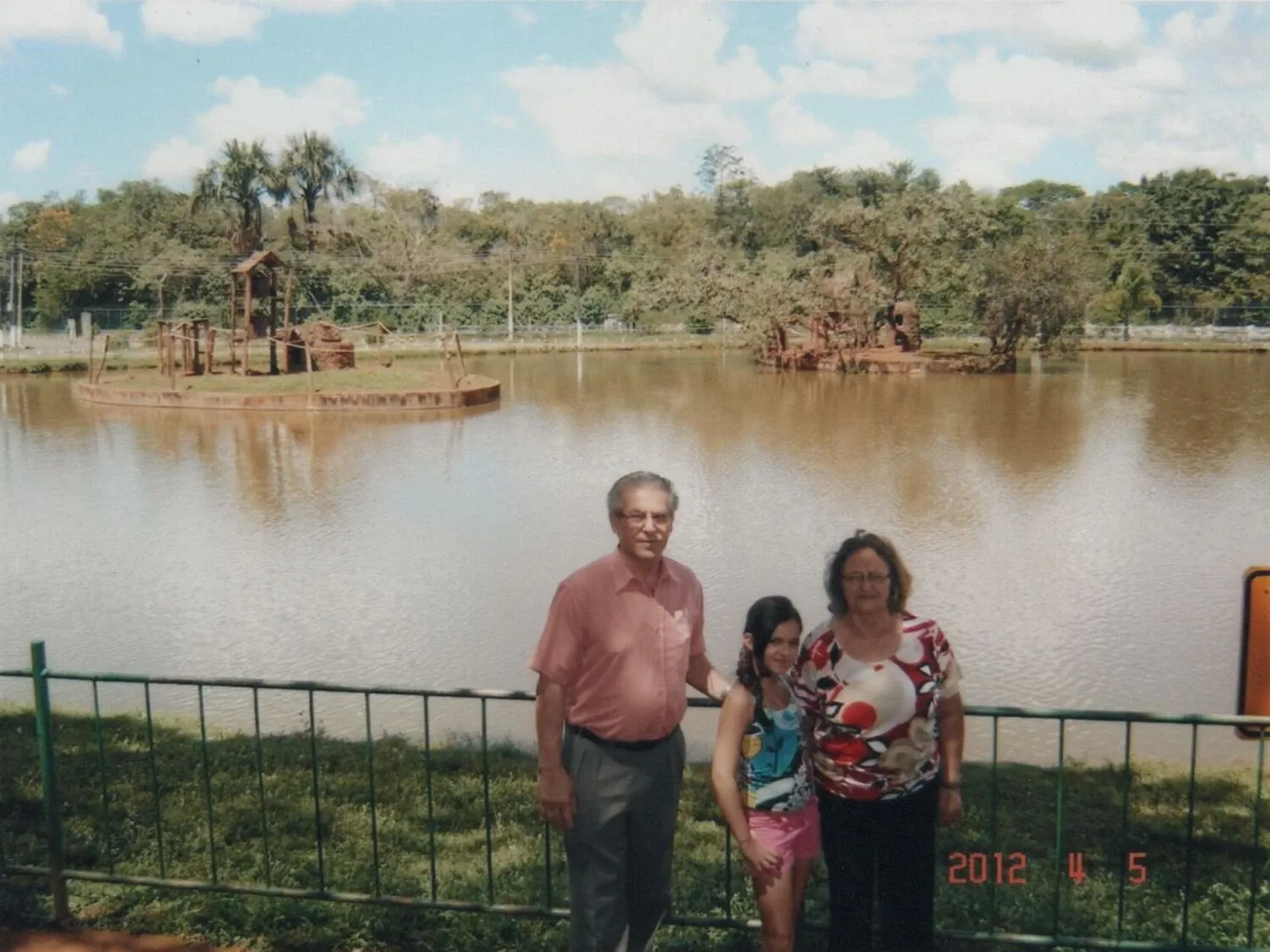Um homem idoso, uma mulher de meia-idade e uma menina de pé na frente de um lago com árvores e algumas estruturas rústicas.