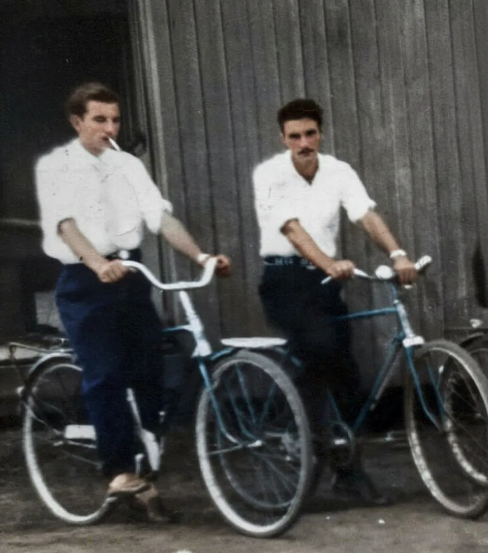 Dois jovens, um com um cigarro na boca, estão de pé ao lado de suas bicicletas, olhando para o lado, em frente a uma parede de madeira.