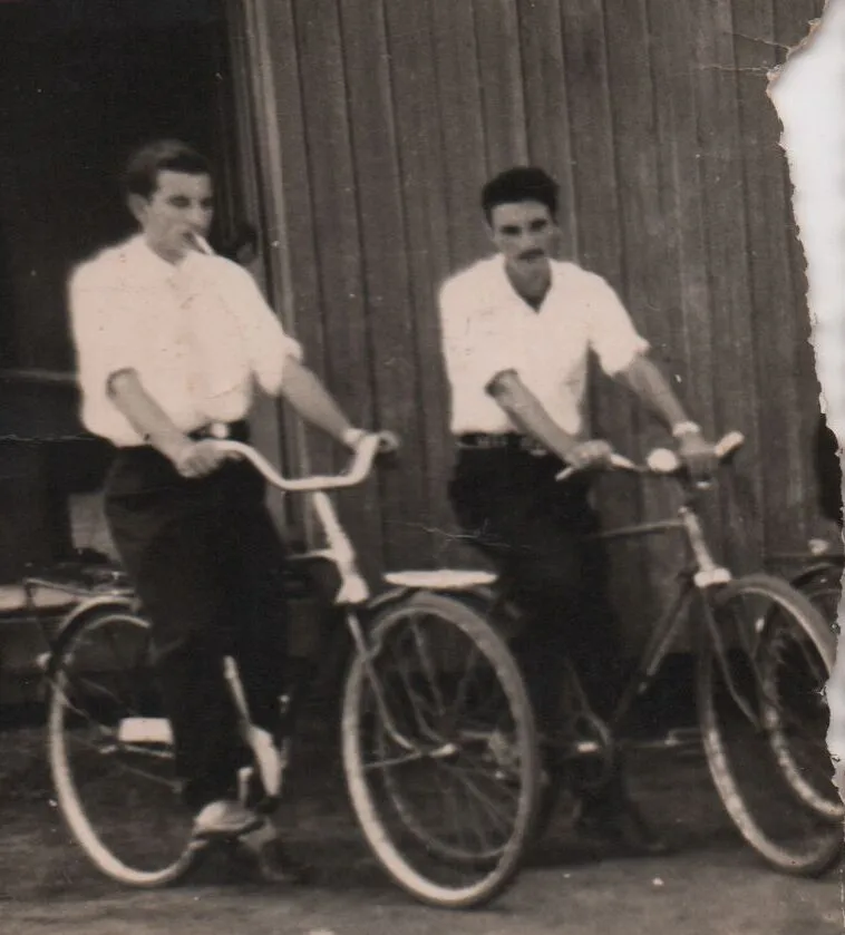Dois jovens, um com um cigarro na boca, estão de pé ao lado de suas bicicletas, olhando para o lado, em frente a uma parede de madeira. (Original)