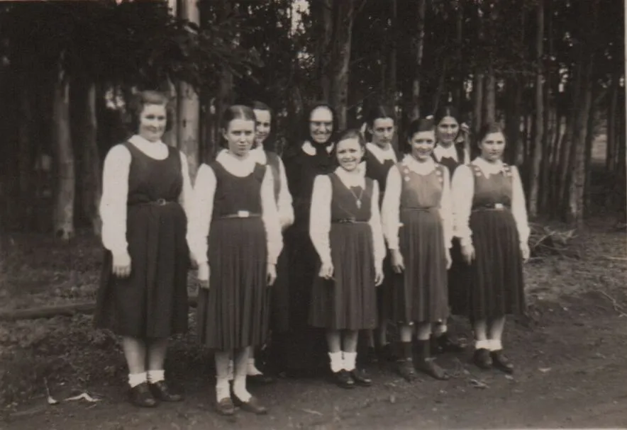 Uma freira e oito jovens estudantes posam para uma foto em pé ao ar livre com um fundo de árvores, em uma imagem em preto e branco com detalhes coloridos. (Original)