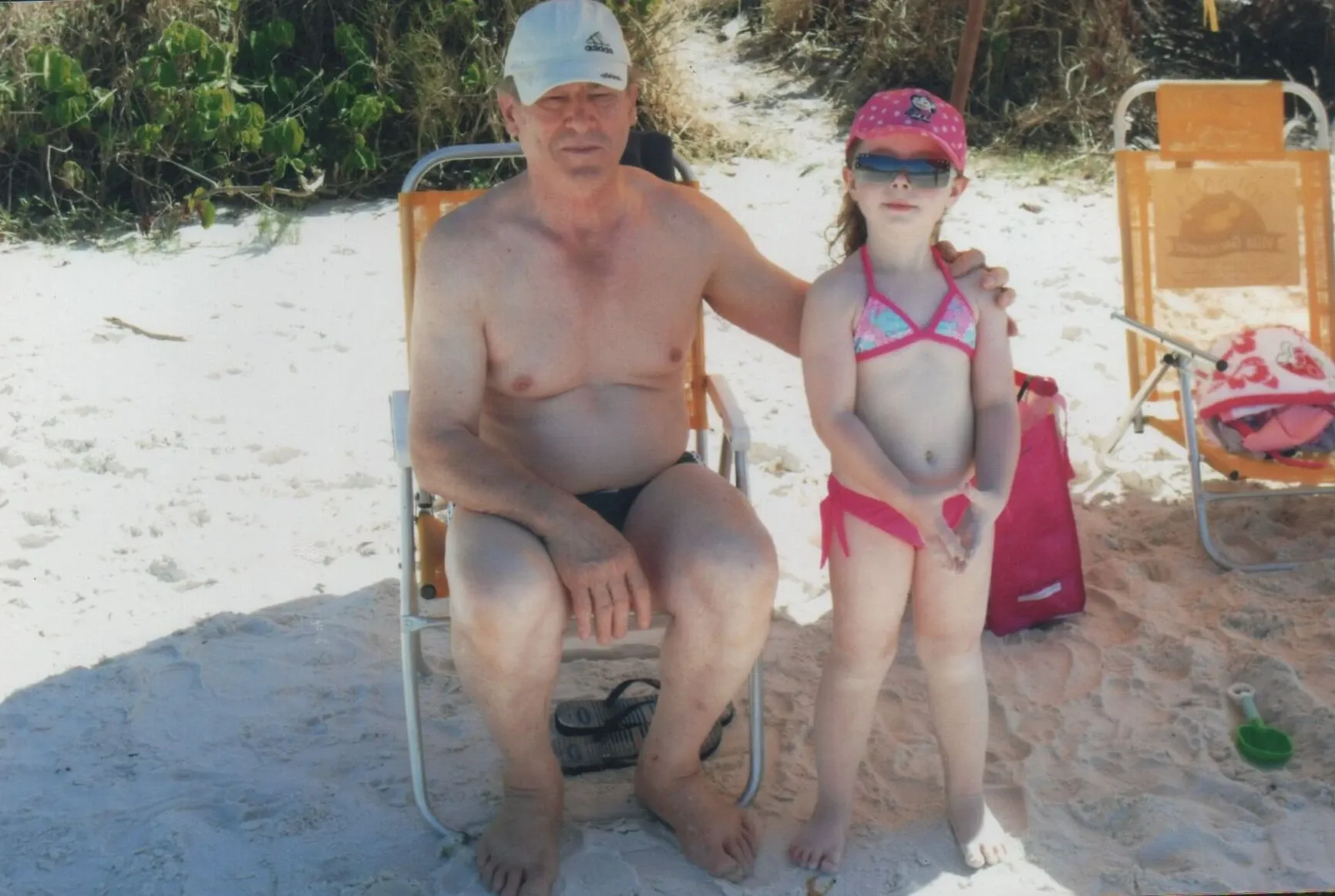 Um homem mais velho de boné e sunga preta sentado em uma cadeira de praia e uma menina de óculos escuros, boné rosa e biquíni rosa, em pé ao lado dele em uma praia arenosa.