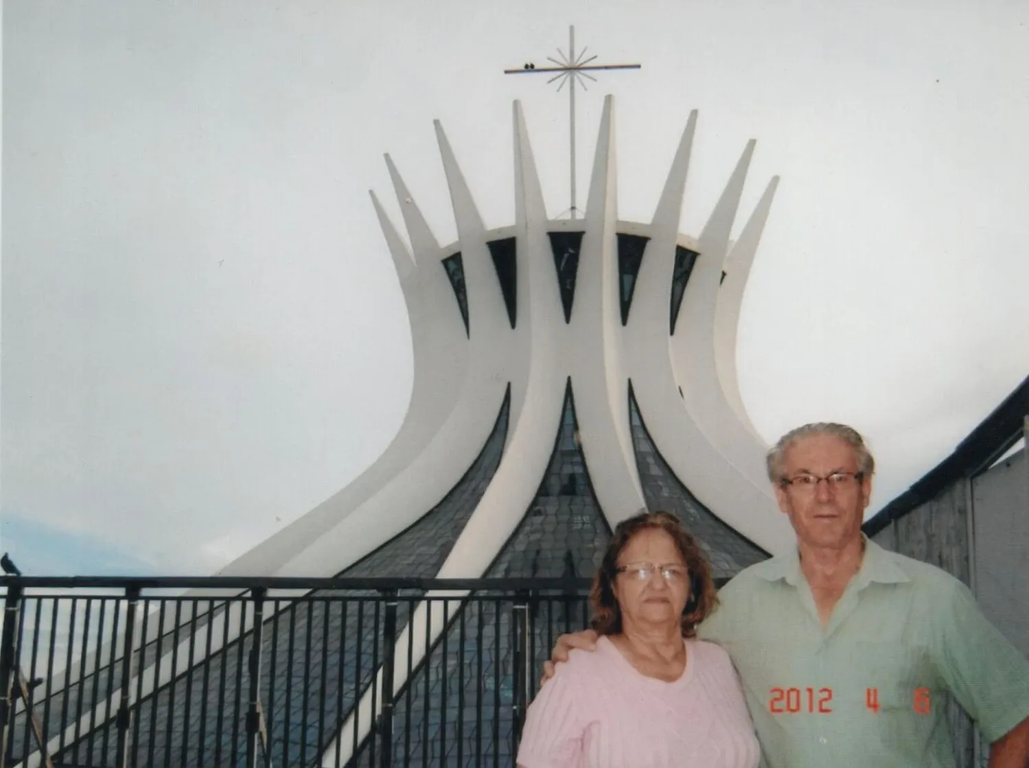 Um homem e uma mulher, provavelmente um casal, estão em pé na frente da Catedral Metropolitana Nossa Senhora Aparecida, no Brasil.