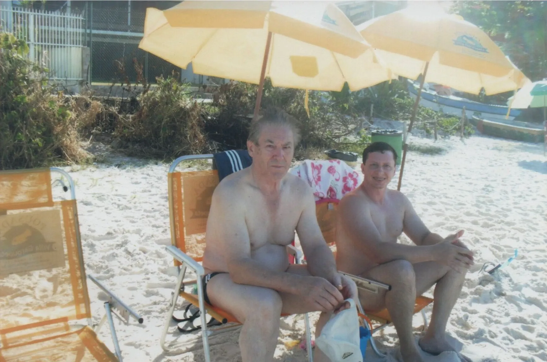 Dois homens sem camisa sentados em cadeiras de praia sob guarda-chuvas de sol em uma praia de areia branca.