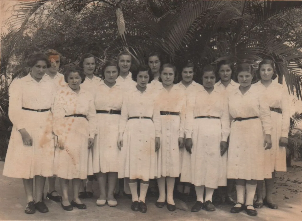Grupo de mulheres jovens em vestidos brancos uniformes, de manga comprida e cinto preto, posando ao ar livre. (Original)