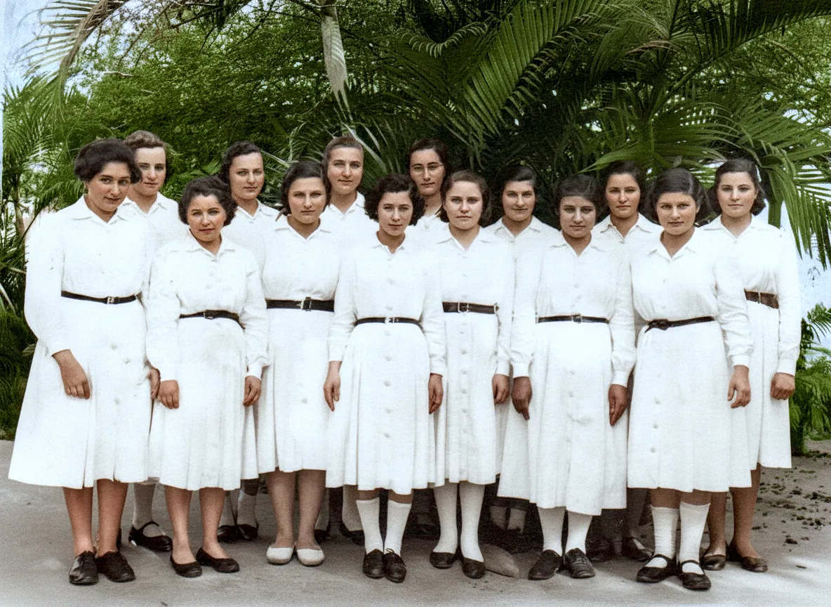 Grupo de mulheres jovens em vestidos brancos uniformes, de manga comprida e cinto preto, posando ao ar livre.