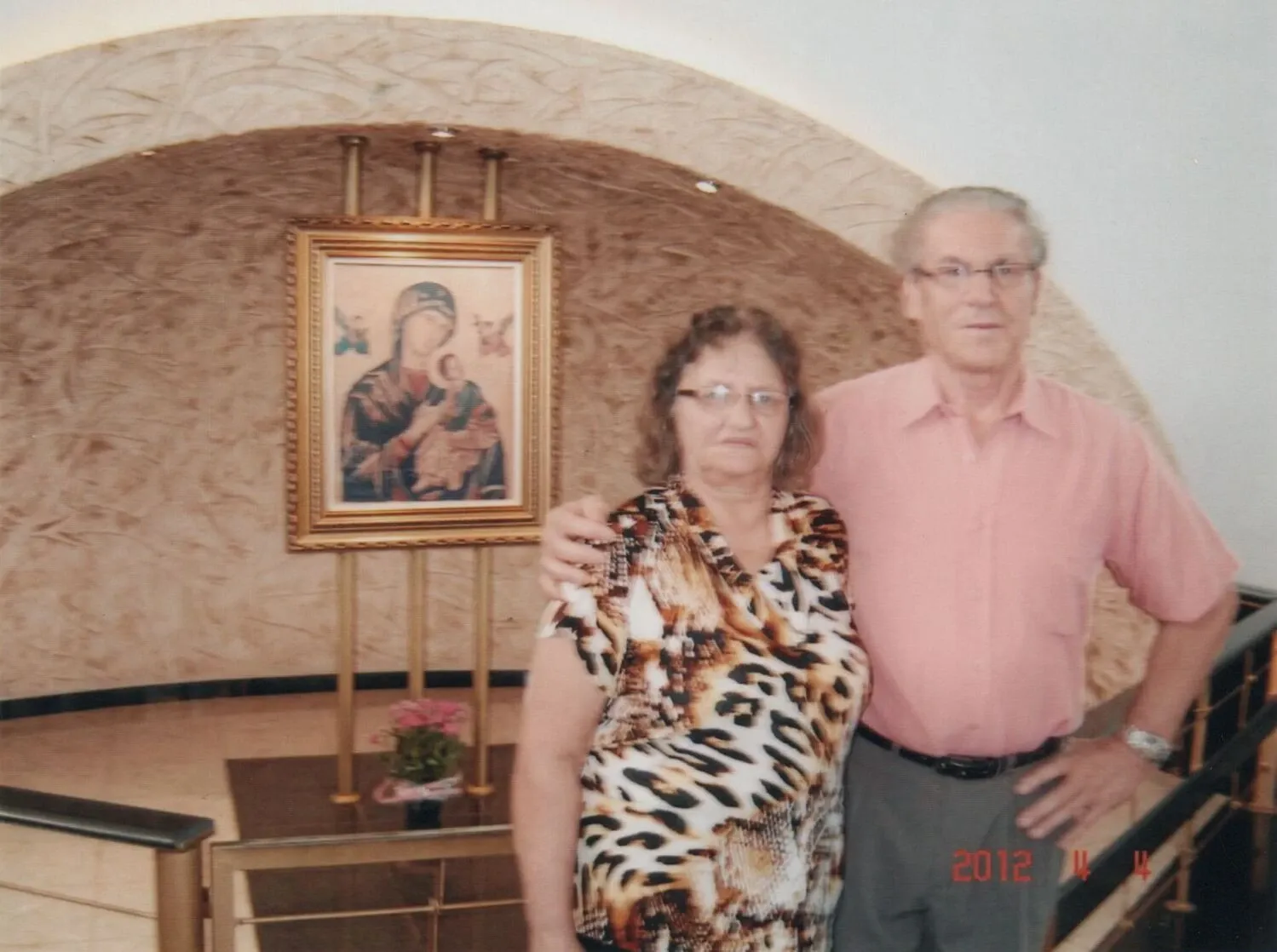 Um casal mais velho está posando na frente de uma pintura de Maria e Jesus em um interior arqueado, possivelmente uma igreja.