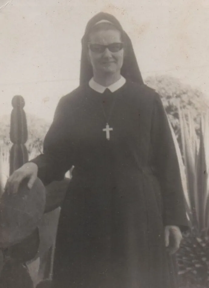 Uma freira sorridente de óculos de sol e hábito azul escuro, com um crucifixo pendurado no pescoço, posa ao ar livre ao lado de um cacto. (Original)