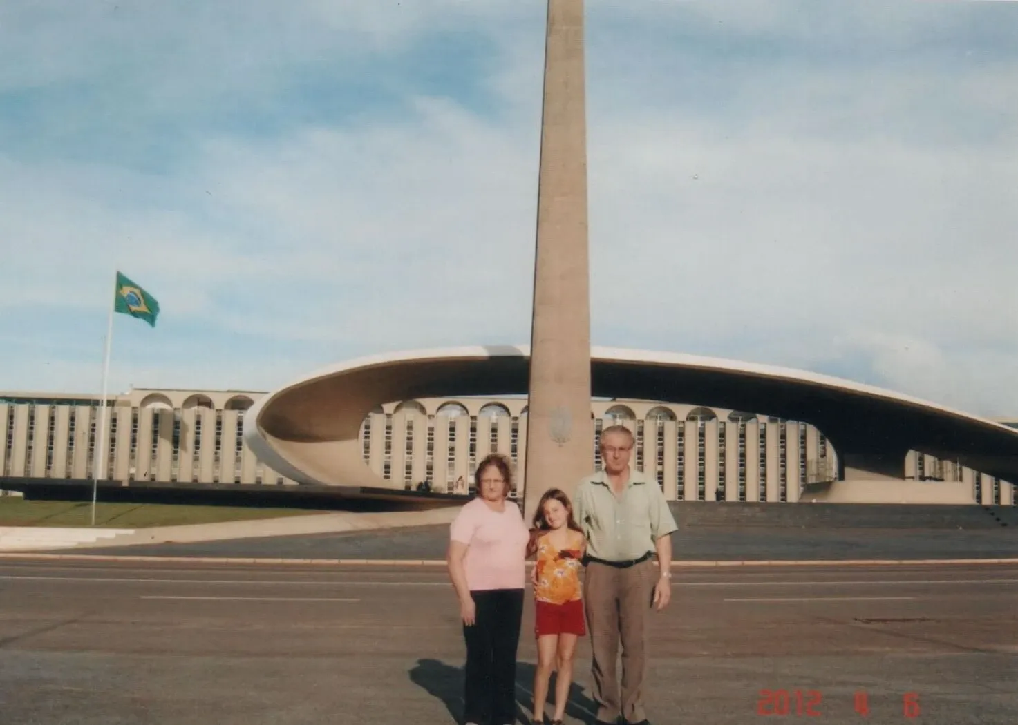Três pessoas, duas mulheres e um homem, estão de pé na frente de um edifício de arquitetura moderna e um obelisco, com a bandeira do Brasil visível ao fundo.