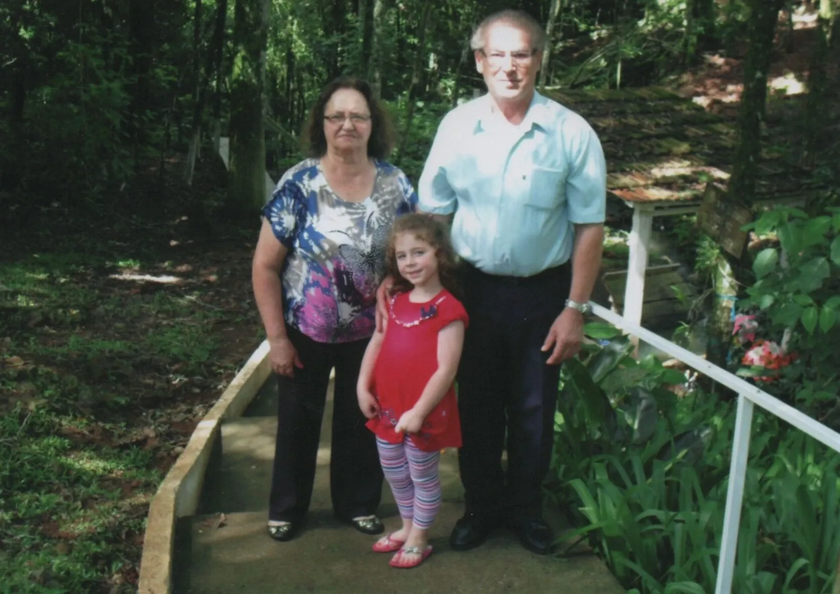 Uma menina em um vestido vermelho e meias listradas cor-de-rosa e brancas fica entre um homem mais velho de camisa azul clara e uma mulher mais velha de blusa estampada, todos em um caminho ao ar livre cercado por folhagens.