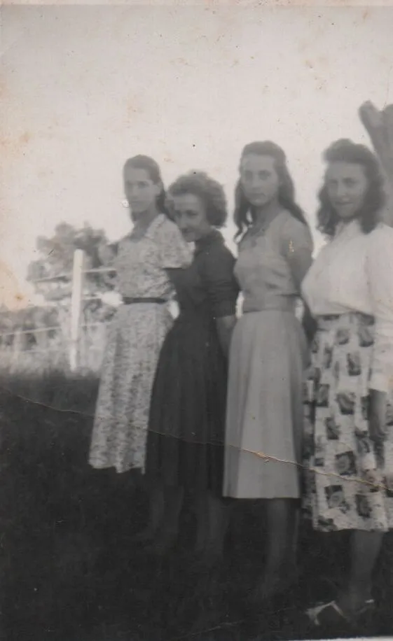 Quatro mulheres jovens em vestidos modestos posam em um campo verde, com uma cerca branca e árvores ao fundo. (Original)