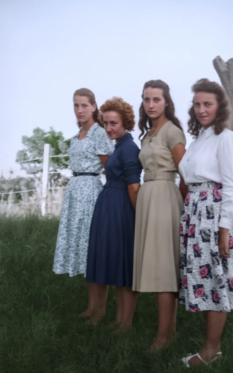 Quatro mulheres jovens em vestidos modestos posam em um campo verde, com uma cerca branca e árvores ao fundo.