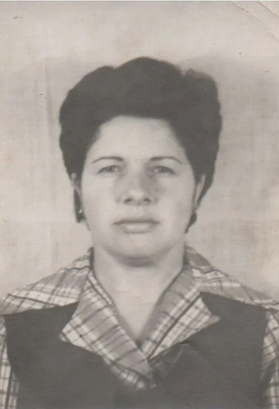 Mulher de meia-idade com cabelo castanho escuro e curto, vestindo uma camisa xadrez azul e branca e um colete azul marinho, olhando diretamente para a frente contra um fundo simples. (Original)